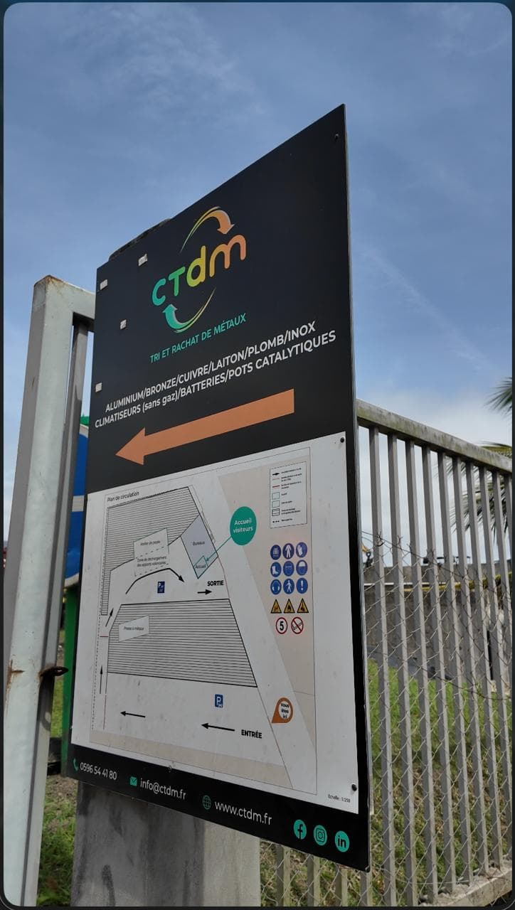 Le panneau d'entrée de CTDM, tri et rachat de métaux, avec le plan de circulation du site, devant un grillage et un ciel bleu.